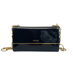 Front view of a Prada flap shoulder bag in shiny black Spazzolato patent leather, featuring a gold-tone Prada logo and a decorative gold-tone chain strap against a white background.