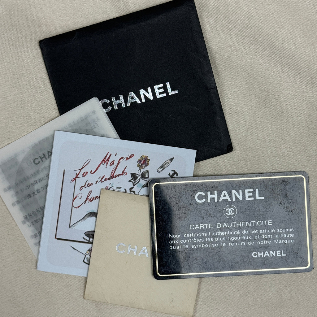 Chanel authenticity documents and packaging laid out on a beige fabric surface, including a black Chanel dust bag, an authenticity card, a receipt, a beige insert card, and a black authenticity card with white text.