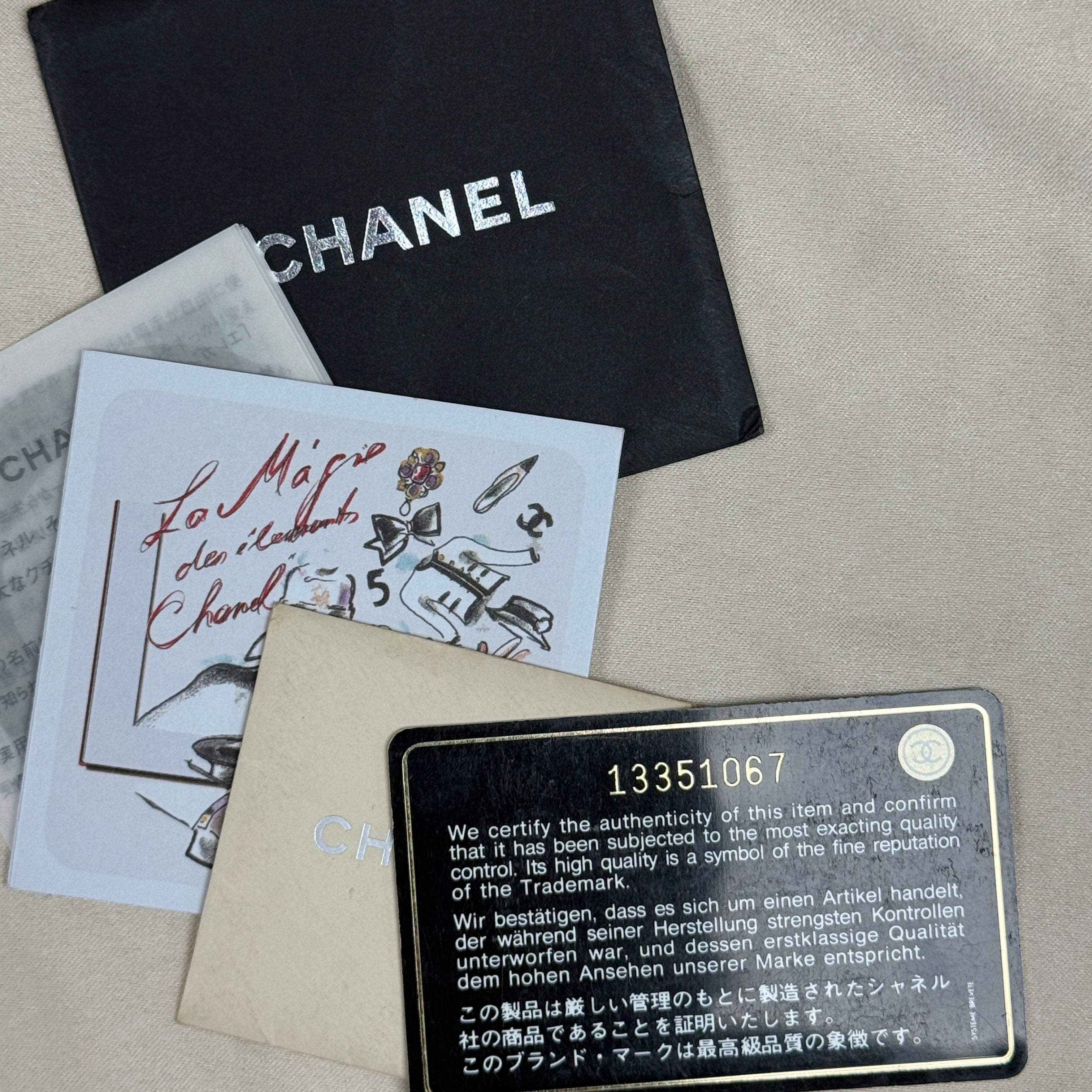 Chanel authenticity card, dust bag, and papers laid out on a beige fabric surface. The black Chanel dust bag is visible at the top, with an authenticity card and a metal plate below it.