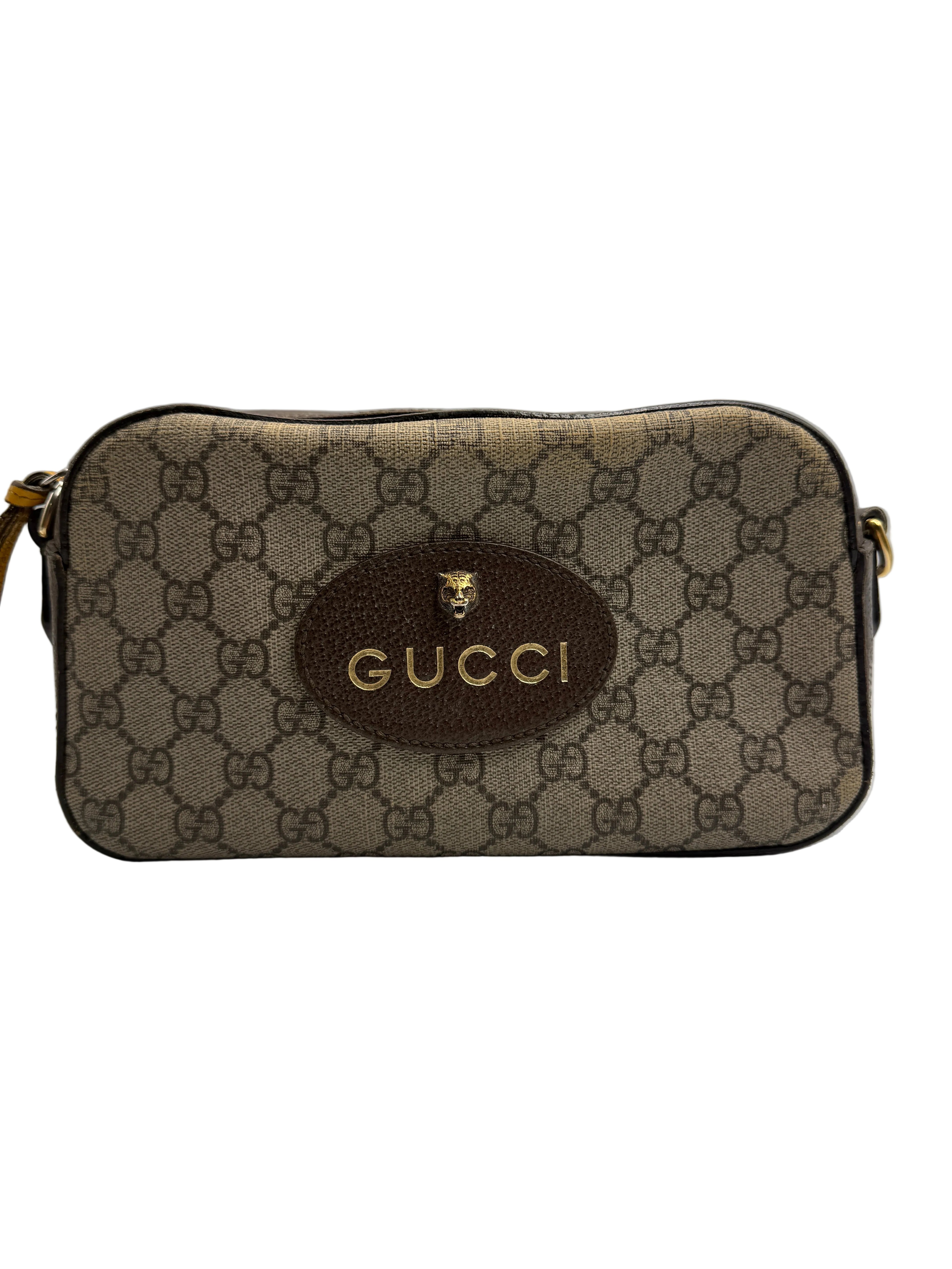 Front view of a Gucci Neo Classic camera bag in GG Supreme canvas with an oval brown leather patch, gold Gucci logo, and feline head hardware.