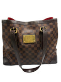 A Louis Vuitton Damier Ebene canvas tote bag with a brown checkerboard pattern, dark brown leather trim, gold-tone hardware, and a red interior lining, shown against a neutral light gray background.