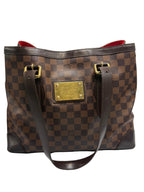 A Louis Vuitton Damier Ebene canvas tote bag with a brown checkerboard pattern, dark brown leather trim, gold-tone hardware, and a red interior lining, shown against a neutral light gray background.