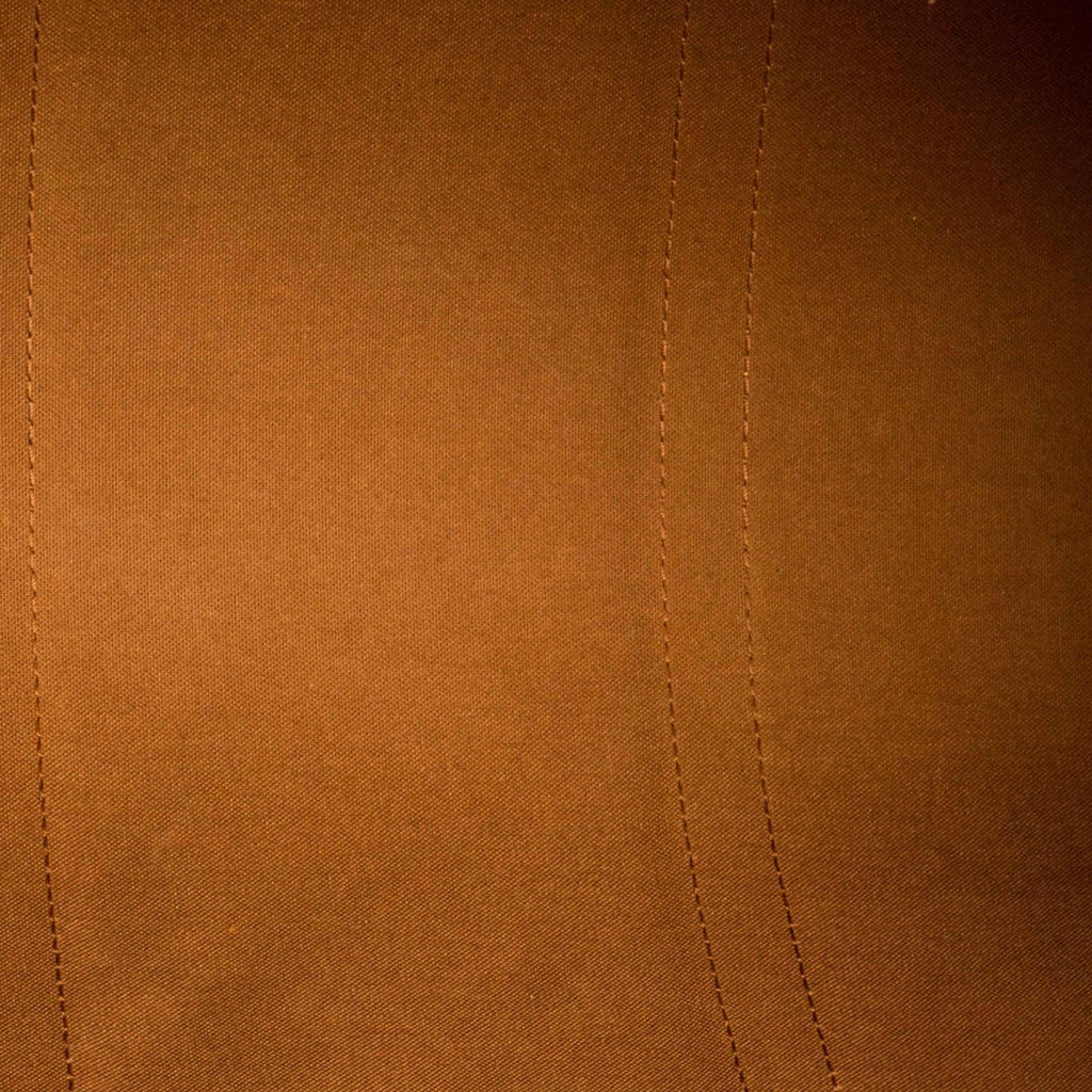 Close-up of a warm brown suede or microfiber fabric surface with visible stitched seam lines and a small portion of a metallic zipper at the top left.