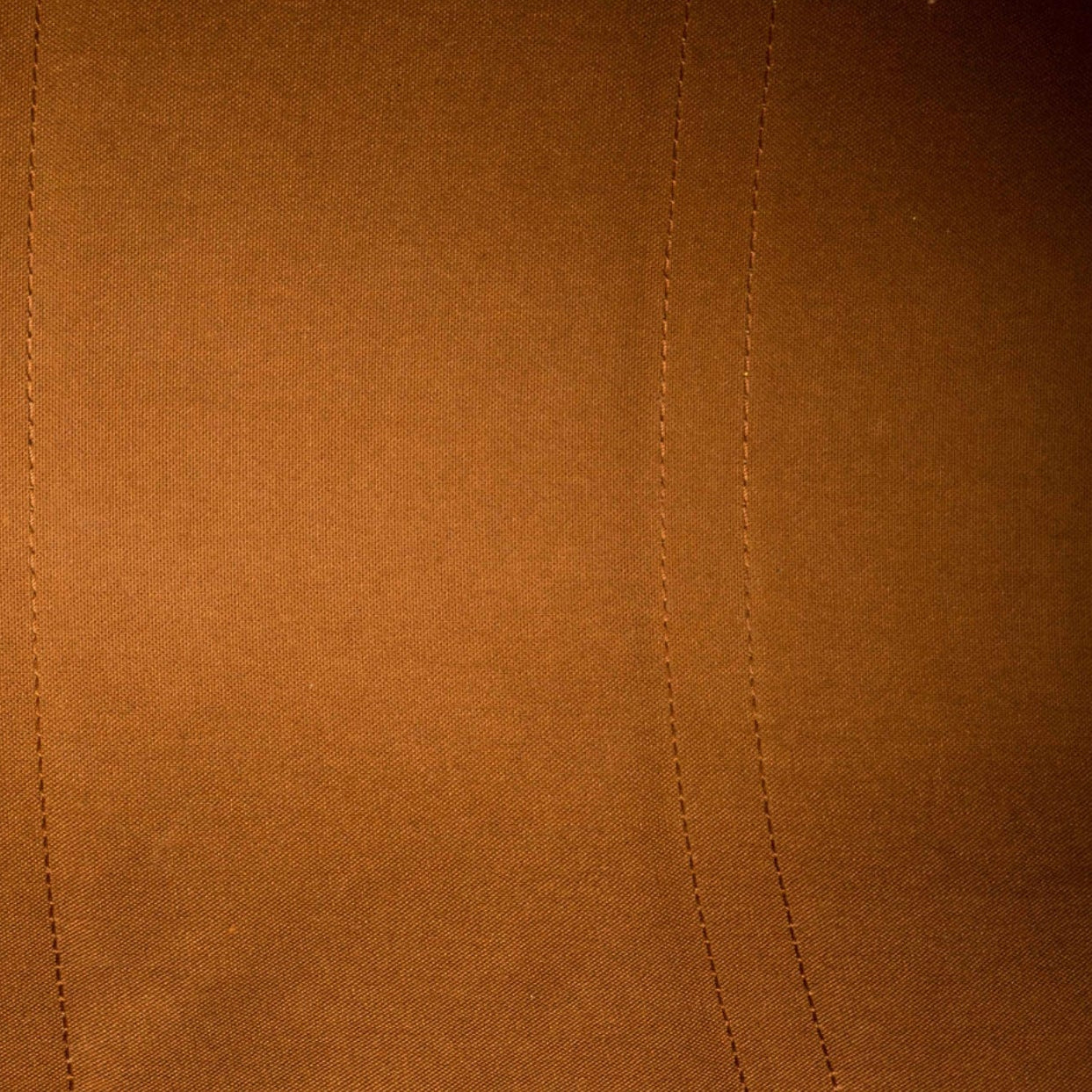 Close-up of a warm brown suede or microfiber fabric surface with visible stitched seam lines and a small portion of a metallic zipper at the top left.