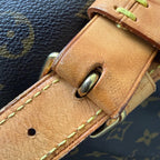 Close-up of a Louis Vuitton monogram canvas bag strap with a tan leather buckle strap, light yellow stitching, and brass hardware.