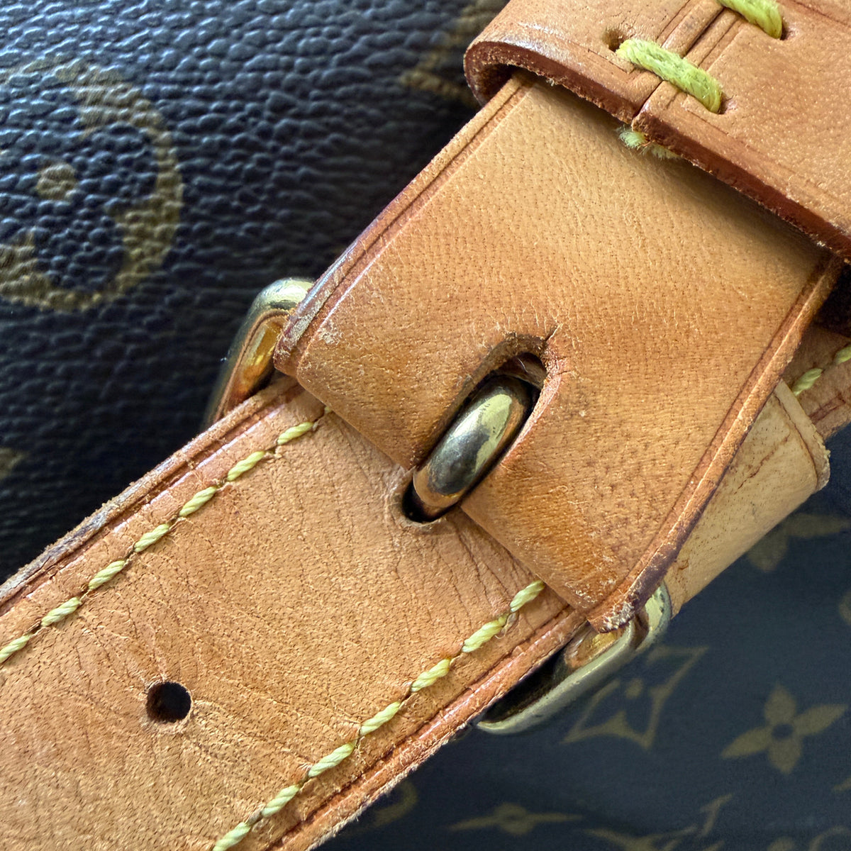 Close-up of a Louis Vuitton monogram canvas bag strap with a tan leather buckle strap, light yellow stitching, and brass hardware.