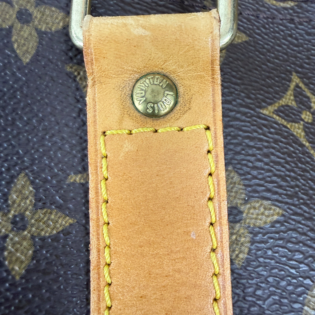 Close-up of a Louis Vuitton monogram bag strap with a tan leather tab, yellow stitching, and a brass rivet stamped with 'LOUIS VUITTON PARIS'.