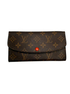 A Louis Vuitton monogram canvas long wallet or card holder with a brown leather texture, tan monogram pattern, and a small red snap button closure, shown on a plain light gray background.