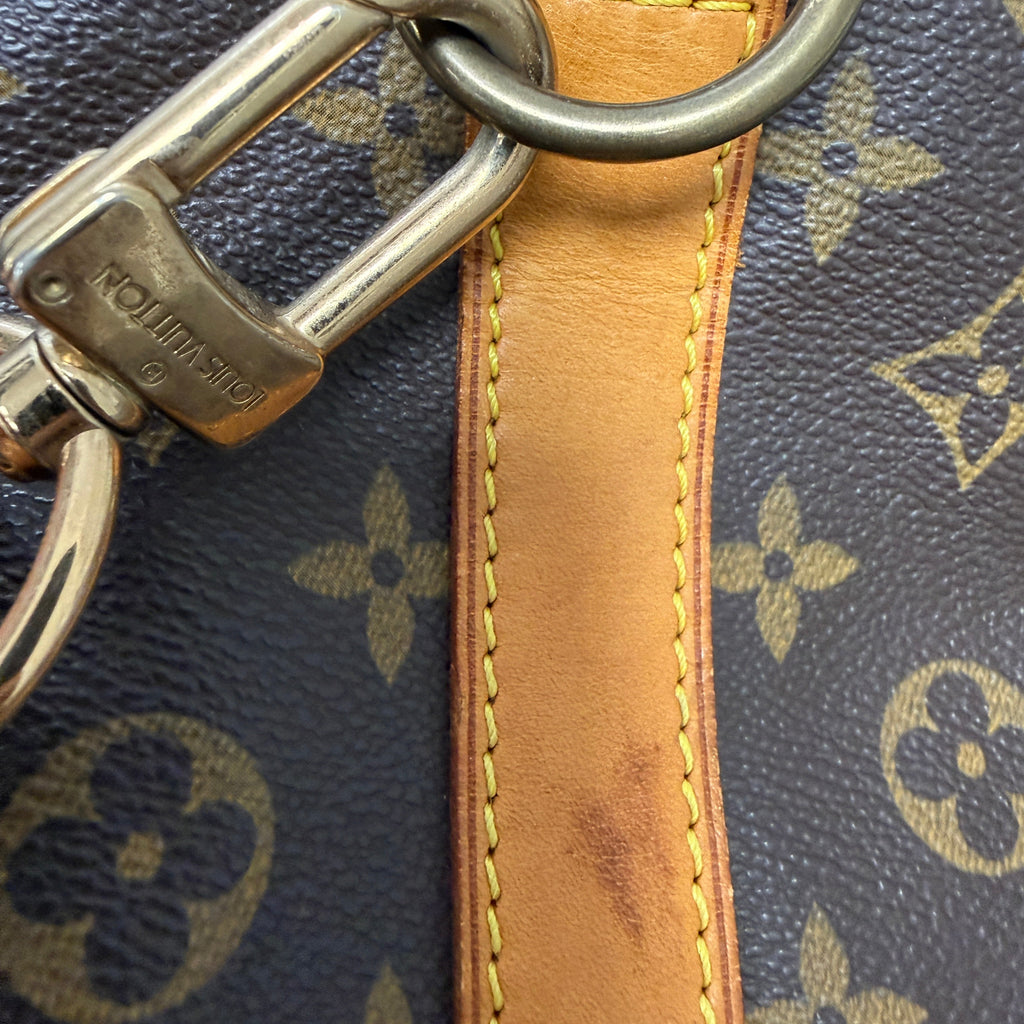 Close-up of a Louis Vuitton monogram bag showing the dark brown coated canvas with tan monogram print, a tan leather strap with yellow stitching, and a silver-tone metal clasp and ring. The Louis Vuitton brand name is embossed on the clasp.